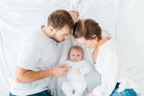 Vista aérea de padres felices acostados en sábanas blancas y mirando al bebé - foto de stock