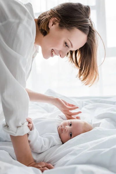 Laughing young mother in white shirt looking at baby on bed — Stock Photo