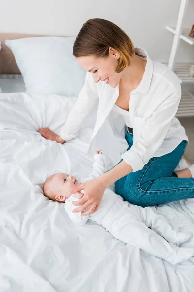 Sorrir jovem mãe em jeans sentado na cama com bebê — Fotografia de Stock