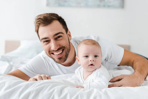 Riendo papá acostado en la cama con lindo bebé y mirando a la cámara - foto de stock