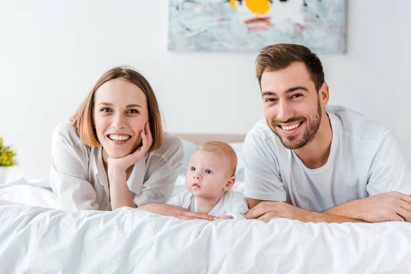 Lächelnde junge Eltern mit Baby, das auf dem Bett liegt und in die Kamera blickt — Stockfoto