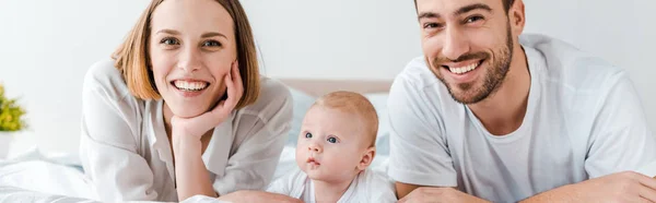 Panoramic shot of smiling young parents with baby lying on bed and looking at camera — Stock Photo