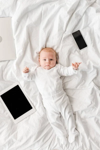 Vista superior del bebé acostado en la cama con computadora portátil, tableta digital y teléfono inteligente con pantalla en blanco - foto de stock