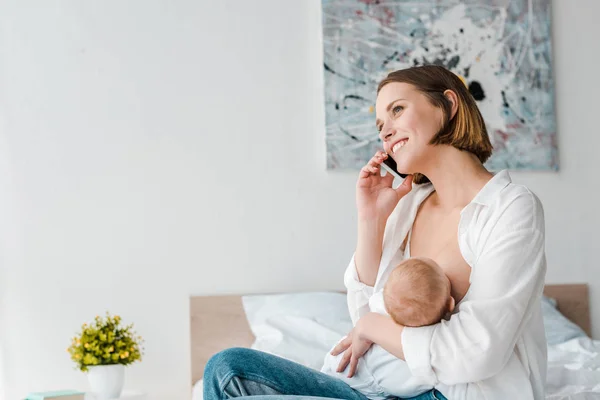 Lächelnde Frau spricht auf Smartphone, während sie ihr Baby zu Hause stillt — Stockfoto