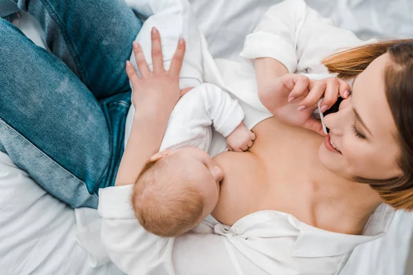 Улыбающаяся женщина разговаривает по смартфону во время кормления грудью ребенка дома — стоковое фото