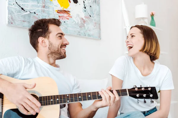 Улыбающийся мужчина сидит на кровати с женой и играет на гитаре — стоковое фото