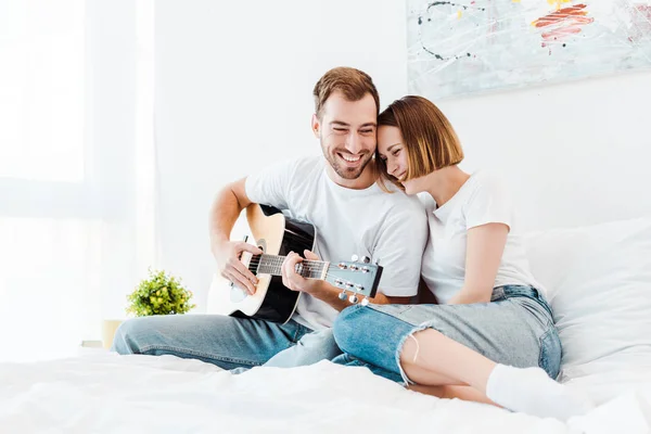 Uomo sorridente seduto sul letto con moglie e suonare la chitarra — Foto stock