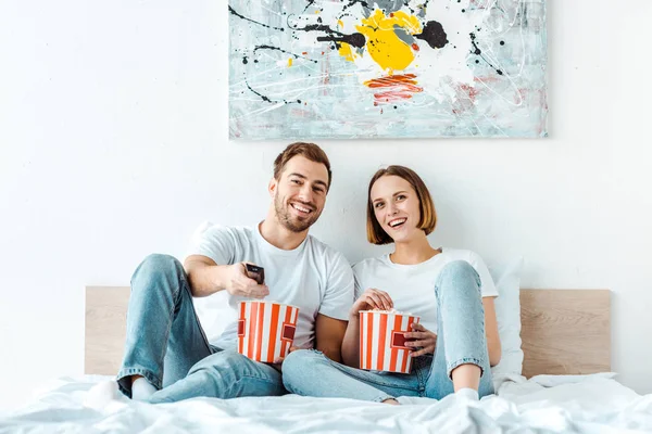 Alegre sonriente pareja comiendo palomitas de maíz en la cama y viendo tv - foto de stock