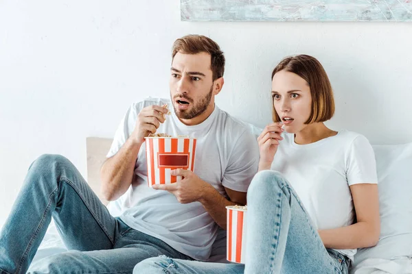 Couple choqué ine jeans assis sur le lit et manger du pop-corn — Photo de stock