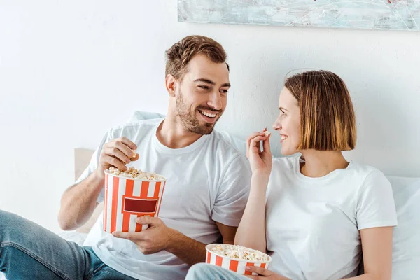 Радісна усміхнена пара їсть попкорн в ліжку і дивиться один на одного — стокове фото