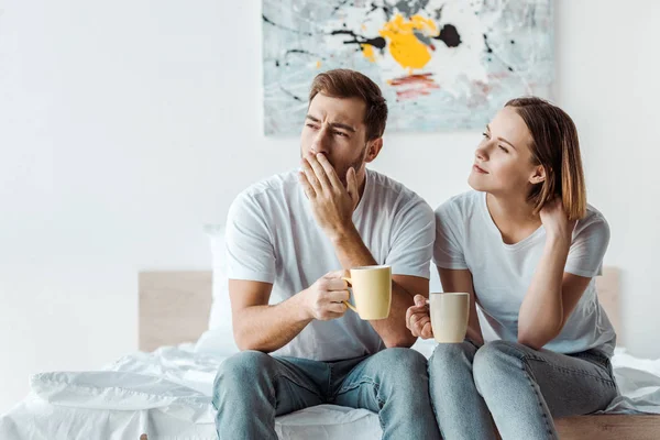 Сонный мужчина держит чашку кофе и закрывает рот рукой, сидя на кровати с женой — стоковое фото