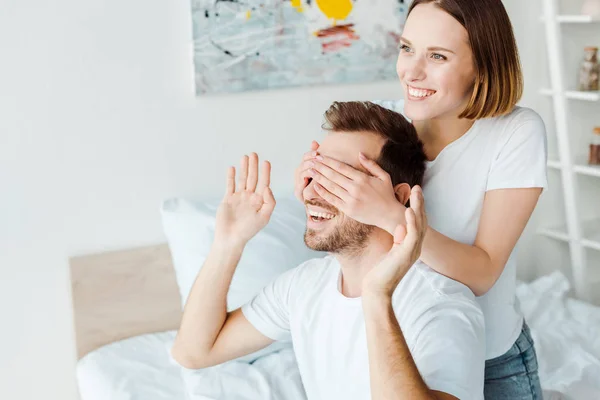 Smiling young woman covering eyes to husband in bedroom — Stock Photo