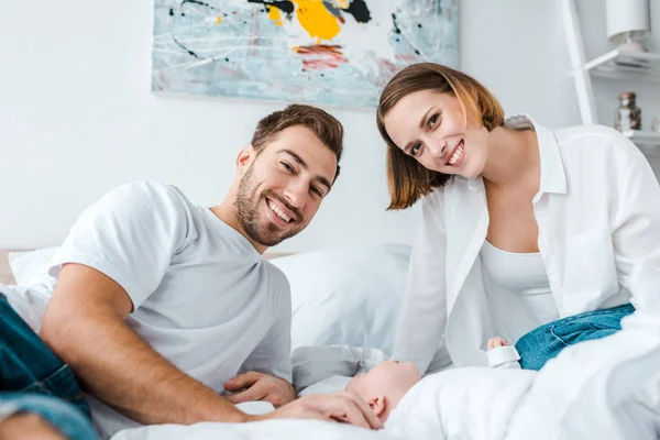 Lächelnde junge Eltern mit Baby, die im Schlafzimmer in die Kamera schauen — Stockfoto