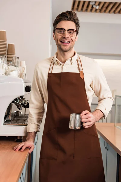 Smiling barista in glasses and brown apron holding milk jug in cafe — Stock Photo