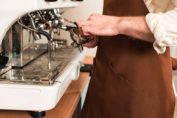 Cropped view of barista in brown apron cleaning portafilter near coffee machine — Stock Photo