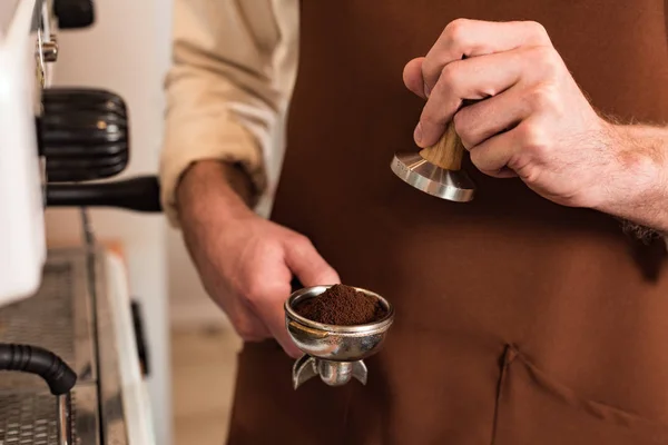 Cropped view of barista in brown apron holding portafilter with ground coffee and tamper — Stock Photo