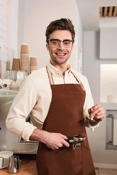 Barista souriant dans des lunettes et tablier marron tenant portafilter avec café moulu et tampon — Photo de stock
