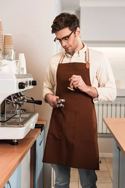 Barista in glasses and brown apron holding portafilter with ground coffee and tamper — Stock Photo
