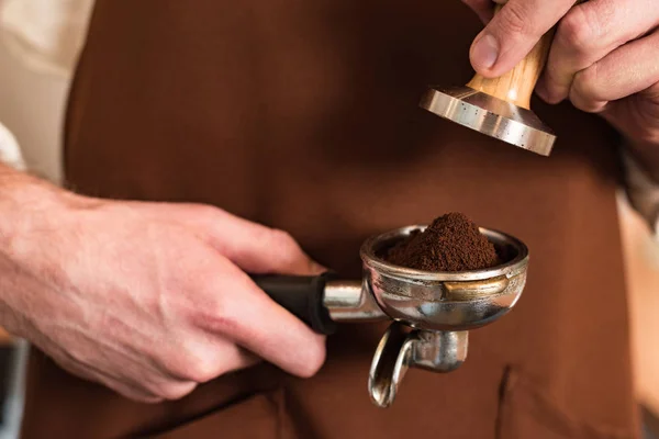 Cropped view of barista in brown apron holding portafilter with ground coffee and tamper — Stock Photo