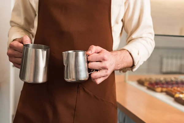 Cropped view of barista in brown apron holding steel milk jugs — Stock Photo