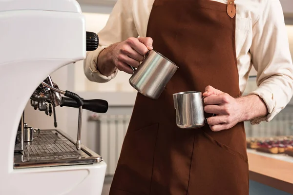 Cropped view of barista in brown apron holding steel jugs — Stock Photo