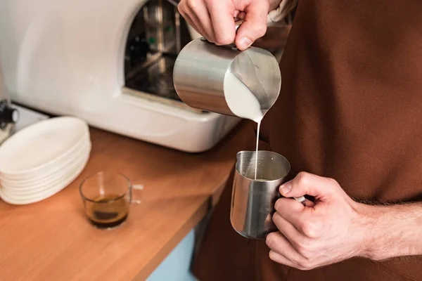 Vista ritagliata di barista in grembiule marrone versando latte in brocca d'acciaio — Foto stock