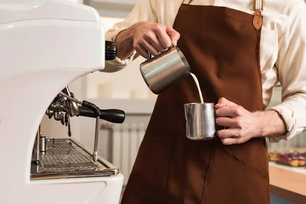 Cropped view of barista in brown apron pouring milk in steel cup — Stock Photo