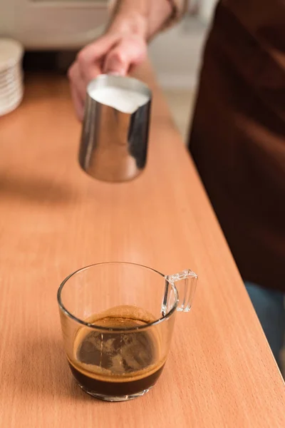Обрезанный вид на баристу с кувшином молока и чашкой кофе на деревянном столе — стоковое фото