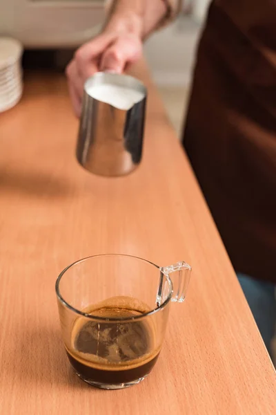 Cropped view of barista with milk jug and cup of coffee on wooden table — Stock Photo