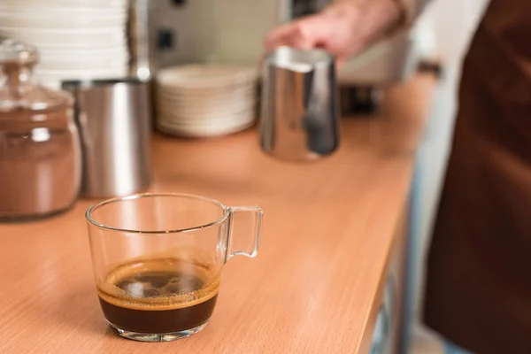 Vista ritagliata di barista con brocca di latte e tazza di caffè sul tavolo di legno — Foto stock