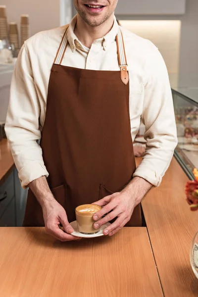 Vue partielle du barista souriant en tablier brun tenant une tasse de café — Photo de stock