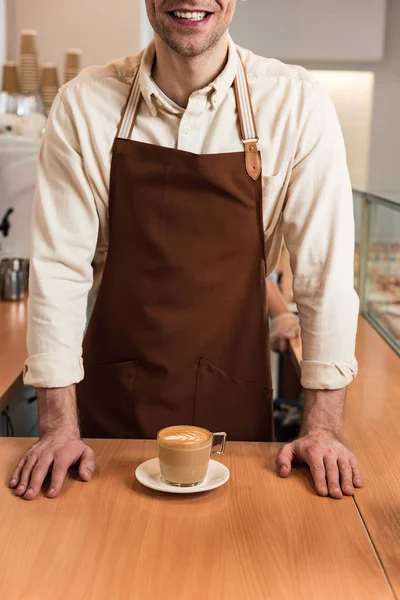 Обрезанный вид смеющегося баристы в коричневом фартуке с чашкой кофе — стоковое фото