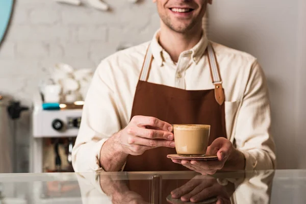 Vista recortada de barista en delantal marrón sosteniendo taza de café con sonrisa - foto de stock