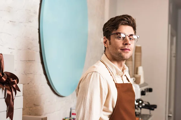 Handsome barista in glasses and uniform looking at camera — Stock Photo