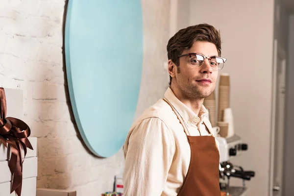 Barista guapo en gafas y uniforme mirando a la cámara - foto de stock