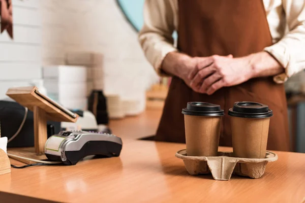 Обрізаний вид бариста в коричневому фартуху і одноразові чашки кави — стокове фото