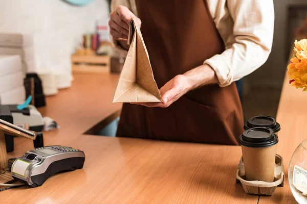 Vista recortada de barista en delantal marrón con bolsa de papel y café para llevar - foto de stock