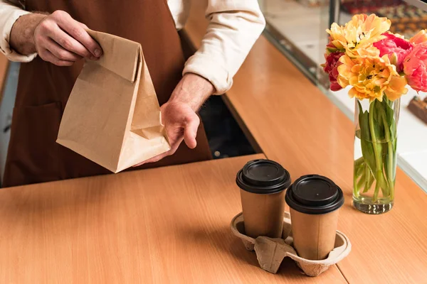 Partial view of barista in brown apron with paper bag and coffee to go — Stock Photo