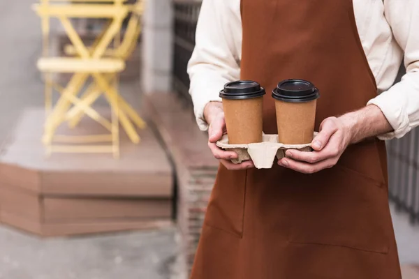 Cropped view of barista in brown apron holding take-out cup carrier with coffee — Stock Photo