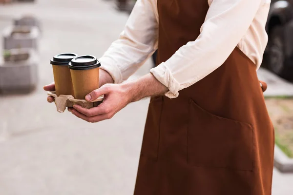 Cropped view of barista in brown apron holding take-out cup carrier with coffee — Stock Photo