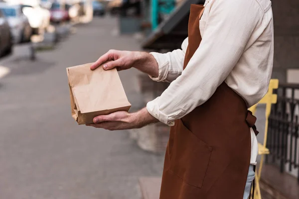 Vista recortada de barista en delantal marrón sosteniendo bolsa de papel en la calle - foto de stock