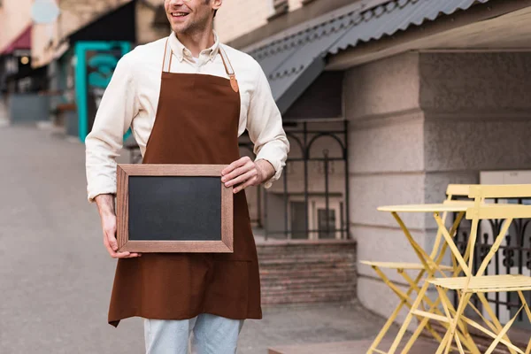 Vista cortada de sorrir barista no avental marrom segurando menu quadro na rua — Fotografia de Stock