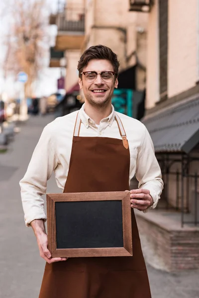Smiling barista in glasses holding chalkboard menu on street — Stock Photo