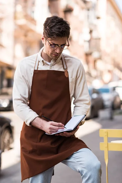 Concentrated barista in glasses and apron writing in notebook on street — Stock Photo