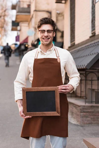 Smiling barista in glasses and apron holding chalkboard menu on street — Stock Photo