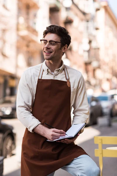 Barista sonriente en gafas y delantal escribiendo en cuaderno en la calle - foto de stock