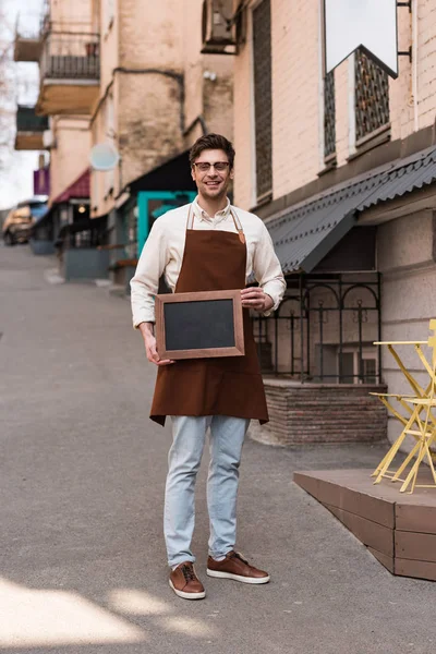 Full length view of smiling barista in glasses and apron holding chalkboard menu on street — Stock Photo