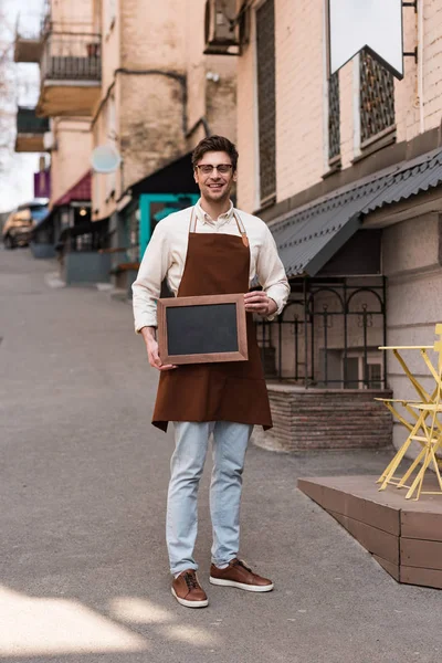 Vue pleine longueur du barista souriant dans des verres et tablier tenant menu tableau noir sur la rue — Photo de stock