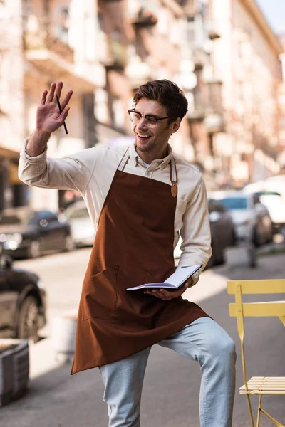 Barista sonriente en gafas y delantal agitando la mano en la calle - foto de stock