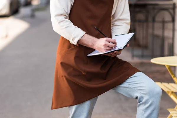 Vue recadrée du barista en tablier marron écrit dans un cahier sur la rue — Photo de stock